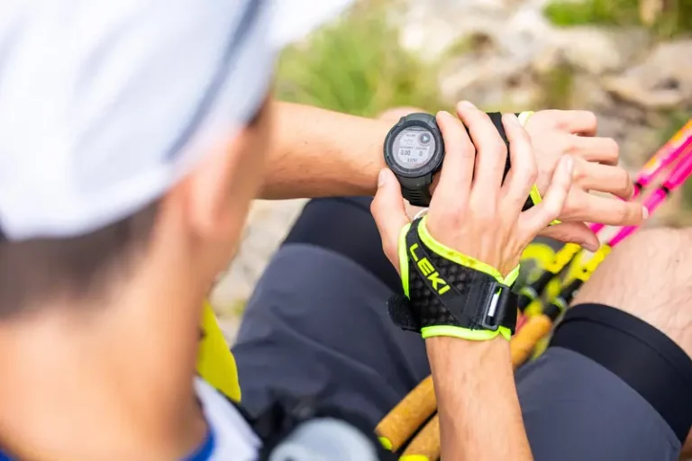 Traileur consultant sa montre GPS avant une sortie en montagne, bâtons de trail à la main